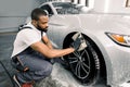 Car washing and detailing photo. African man worker in protective overalls and rubber gloves, washing car wheel rims of