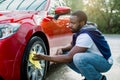 Car washing on open air. Young hipster African bearded man cleaning a wheel, car rims of modern luxury red car with Royalty Free Stock Photo