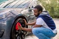 Car washing on open air. Young hipster African bearded man cleaning a wheel, car rims of modern luxury gray electric car Royalty Free Stock Photo