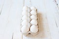 A cardboard box with a dozen white chicken eggs lying on a white painted wooden surface. Background for livestock products