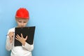 boy in a protective construction helmet concentrating on writing something with a pen on a tablet on a blue background