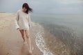 Carefree beautiful woman in knitted sweater and with windy hair running on sandy beach at cold sea waves, having fun. Stylish Royalty Free Stock Photo