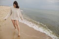 Carefree beautiful woman in knitted sweater and with windy hair running on sandy beach at cold sea waves, having fun. Stylish Royalty Free Stock Photo