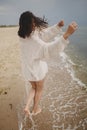 Carefree beautiful woman with windy hair running on sandy beach at cold sea waves, having fun. Stylish young happy female in Royalty Free Stock Photo