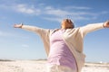 Carefree biracial senior woman with arms outstretched enjoying sunny day at beach against blue sky