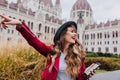 Carefree caucasian girl in trendy red jacket fooling around during european trip. Excited young lady in hat exploring Royalty Free Stock Photo