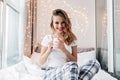 Carefree girl with cheerful smile posing in bed in her room. Indoor portrait of debonair blonde woman holding cup of tea Royalty Free Stock Photo