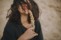 Carefree hipster girl with windy hair sitting and smiling on sandy beach, holding herb. Stylish tanned young woman in modern