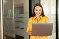 Carefree inspired female employee standing in modern office space and working laptop, cheerful businesswoman in yellow