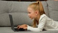 Carefree little kid pretty blonde girl child daughter using laptop lying on grey sofa surfing internet chatting with Royalty Free Stock Photo