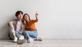 Carefree millennial Asian man and woman sitting on floor near white studio wall and pointing at copy space, banner Royalty Free Stock Photo