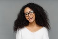 Carefree overjoyed dark skinned woman laughs happily, being in high spirit, tilts head, has curly dark hair, wears white jumper, Royalty Free Stock Photo