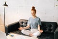 Carefree redhead young woman takes deep breath, practices yoga or meditation, sitting on sot couch