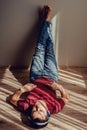 Carefree relaxed man in jeans, wine red t-shirt listening to music lying down on parquet floor, feet on wall. Natural light and Royalty Free Stock Photo