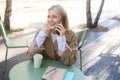 Carefree young woman in trench, talking on mobile phone and smiling, sitting outside cafe and drinking coffee, answer a