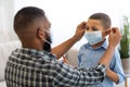 Careful African Father Putting Face Mask On Son Indoors