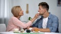 Careful mother wiping adult sons mouth during having tea at home, overprotection