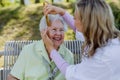 Caregiver helping senior woman to comb hair and make hairstyle when sitting on bench in park in summer. Royalty Free Stock Photo