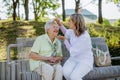 Caregiver helping senior woman to comb hair and make hairstyle when sitting on bench in park in summer. Royalty Free Stock Photo