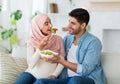 Caring arab man giving his young pregnant wife fresh vegetable salad, sitting on sofa in living room at home Royalty Free Stock Photo