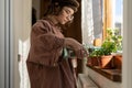 Caring creative teenage girl watering houseplant from bottle on wooden windowsill. Home help parents