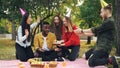 Caring friends are bringing cake to African American man sitting on blanket in park on picnic with closed eyes, he is
