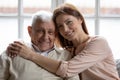 Caring grownup granddaughter hugs elderly 75s grandfather seated on sofa