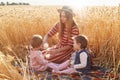 Caring helpful mother sitting on blanket at wheat field with her lottle daughters, filling glasses with milk from bottle, wearing