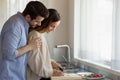 Caring millennial husband hugging shoulders of beloved wife preparing breakfast