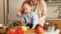 Caring mother giving fruit puree on spoon to her baby son playing with vegetables and cookware on kitchen Royalty Free Stock Photo