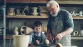 Caring silver-haired grandfather is teaching young cute grandson to work with clay on throwing-wheel in small workshop
