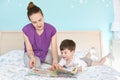 Caring young mother reads magazine with pictures for children to her small son, pose together at bed against cozy interior and