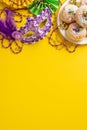 Vertical top view of plate piled with scrumptious donuts, luxurious masquerade mask, colorful bead necklaces on yellow setting