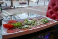 Carpaccio of red beef and pork with tomatoes sprinkled with cheese and greens on a white rectangular plate on a table in a cafe Royalty Free Stock Photo