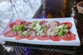 Carpaccio of red beef and pork with tomatoes sprinkled with cheese and greens on a white rectangular plate on a table in a cafe Royalty Free Stock Photo