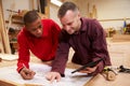 Carpenter With Apprentice Looking At Plans In Workshop