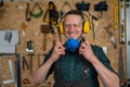 A carpenter in goggles and earmuffs removes a protective respirator.