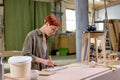 Carpenter woman paints wooden slat for production of furniture on table in workshop