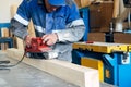 Carpenter in work clothes processes wood with grinder. Polishing of wood surface. Middle-aged man works at workbench in Royalty Free Stock Photo