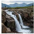 A cascading waterfall flows over rugged, uneven rocks, surrounded by lush greenery.