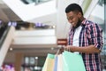 Cashback Bonus. Happy African Man Holding Shopping Bags And Smartphone In Mall