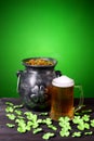 Cast iron pot with four-petal lucky shamrock leaf, mug of beer, clover leaves on dark wooden table and green gradient background. Royalty Free Stock Photo