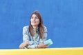 Casual adult woman lean on yellow wall holding a mobile phone while looking away outdoors against a blue background Royalty Free Stock Photo