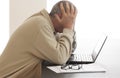 Casual dressed man is bending his head over computer in despair while he hides his head in his hands. Pair of glasses lying on a p Royalty Free Stock Photo