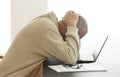 Casual dressed man bends his head over computer in despair while he hides his head in his hands. Pair of glasses lying on a pile o Royalty Free Stock Photo