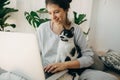 Casual girl working on laptop with her cat, sitting together in modern room with pillows and plants. Home office. Young woman Royalty Free Stock Photo