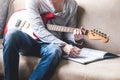 Casual young man playing guitar and writing some data in notebook on sofa at home. Royalty Free Stock Photo