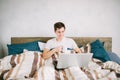 Casual young man using laptop in bed at home and drinking coffee Royalty Free Stock Photo