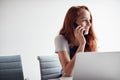 Casually Dressed Young Businesswoman Working On Laptop At Desk In Modern Workplace Making Phone Call Royalty Free Stock Photo