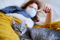 Cat is lying at the feet of a woman with coronavirus. Redhead girl looks at the thermometer lying in bed